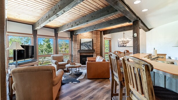 A spacious living area with a fireplace, wooden beams, and large windows offering a view of the outdoors. - Walk to Gondola | Cozy 2BR Condo | Pool, Hot Tubs and Fireplace! (Steamboat Springs)