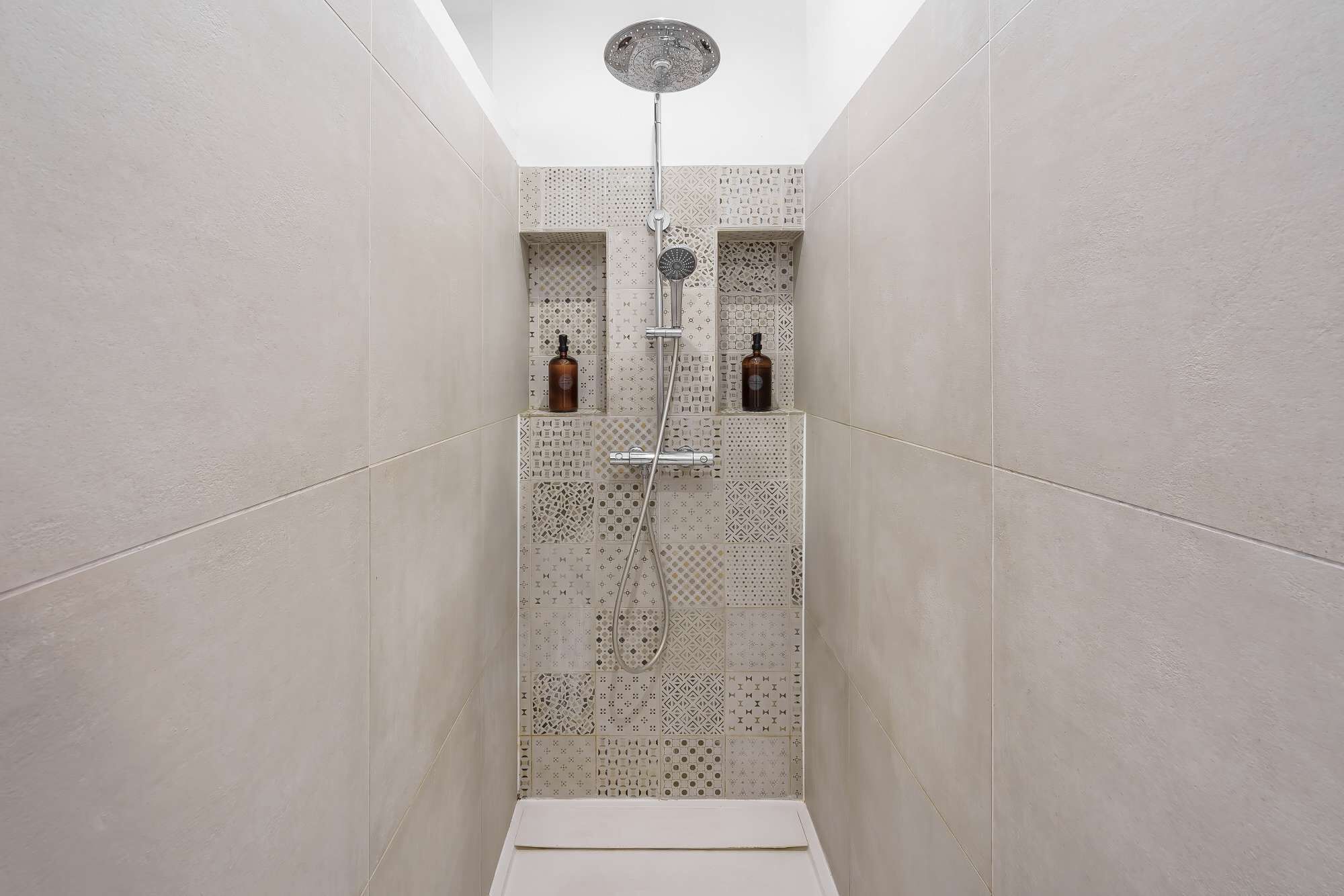 A narrow shower with patterned tiles, a ceiling-mounted showerhead, and two bottles of liquid soap on the wall.