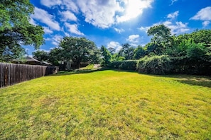 A well-maintained grassy backyard with a wooden fence, surrounded by lush greenery and trees under a clear blue sky.