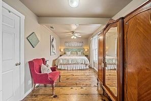 A bedroom with a wooden floor, a bed with a floral bedspread, a red chair, a ceiling fan, and a wooden wardrobe.