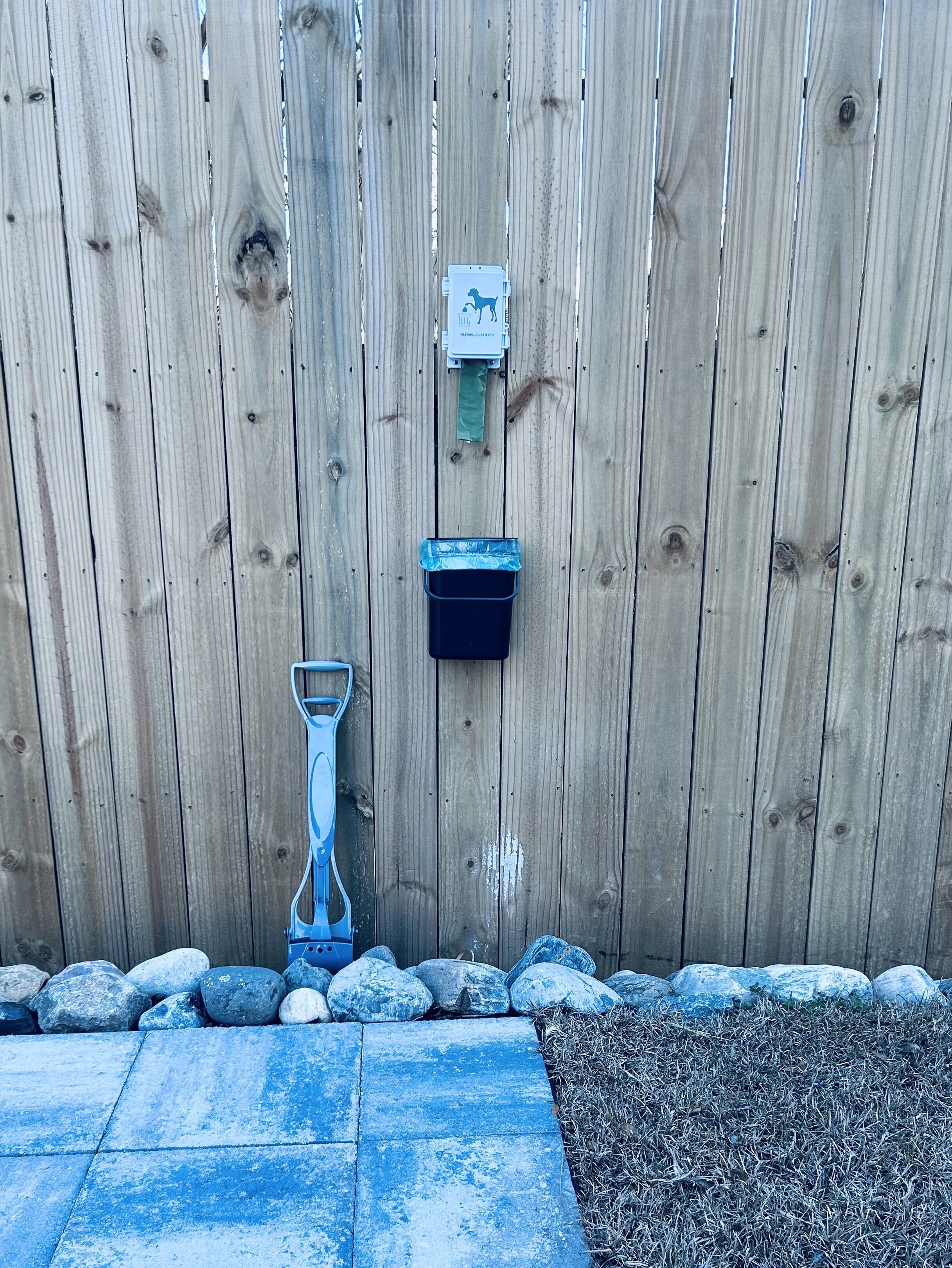 Een houten hek met een metalen poort, een blauw metalen werktuig, en een zwarte vuilnisbak.