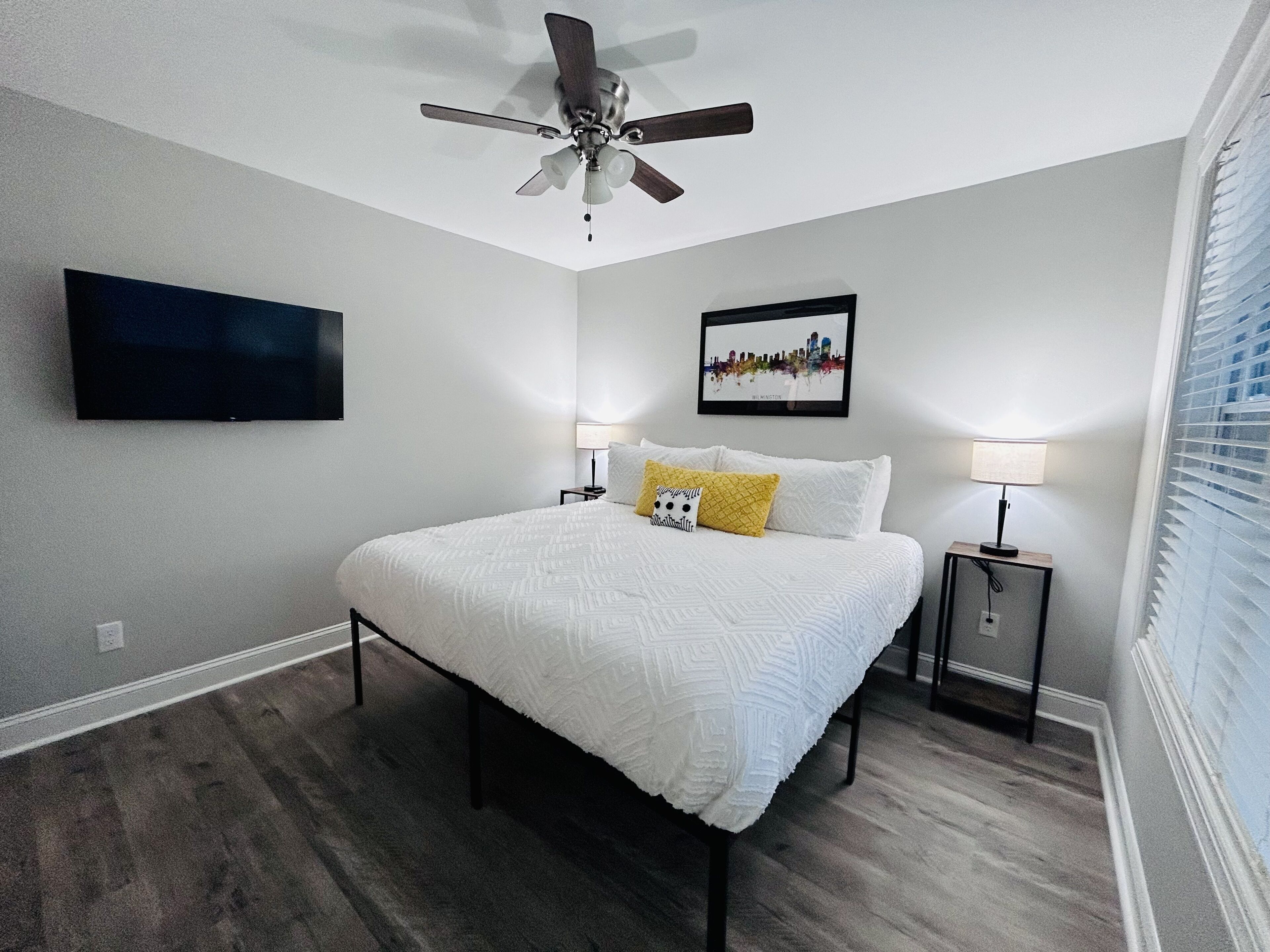 Een slaapkamer met een bed, een plafondventilator, een aan de muur gemonteerde televisie, een stadsgezicht in een lijst en een nachtkastje met een lamp.
