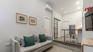 A modern living room with a grey sofa, two green cushions, and a small rug. There is a kitchen area with a fridge, a sink, and a window. - Waou Fresh Confort I (Madrid)