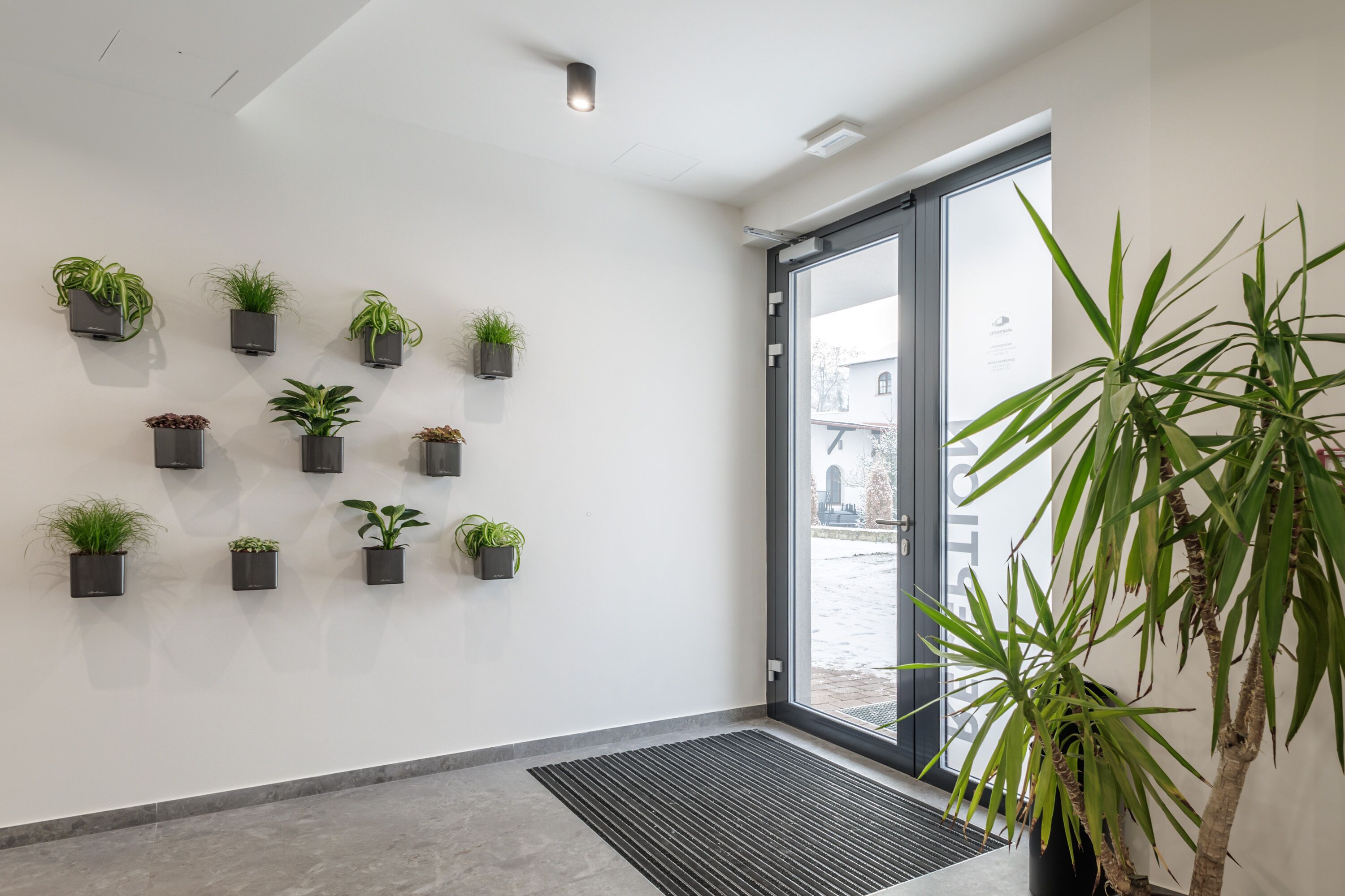 A modern interior with a series of potted plants on the wall, a large window with a view of the outside, and a black floor mat near the entrance.