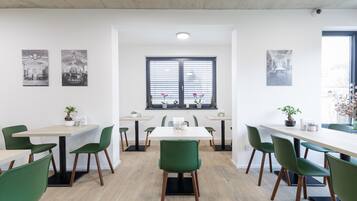 Un comedor moderno con varias mesas y sillas verdes, que cuenta con un amplio ventanal con persianas y plantas en macetas.