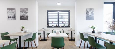 A modern dining area with multiple tables and green chairs, featuring a large window with blinds and potted plants.