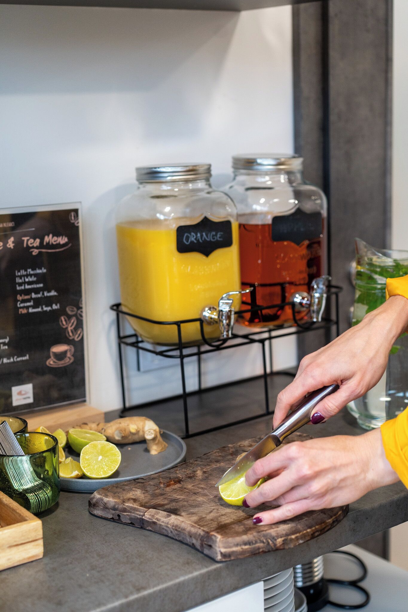 Una encimera de cocina con una persona cortando un limón, un dispensador de zumo con naranja y otra bebida, y un tablero de menú que ofrece opciones de té y café.