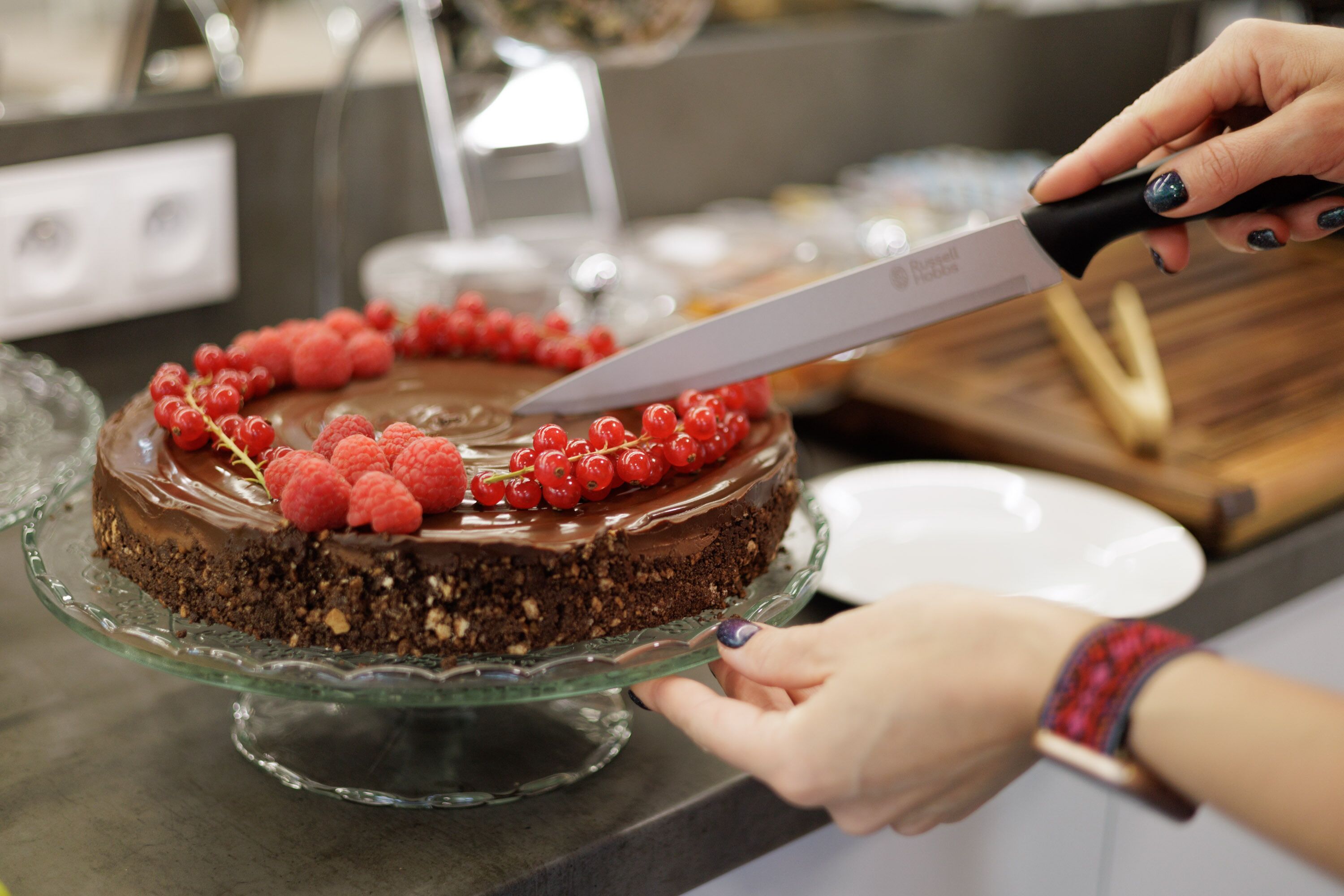 A person cutting a chocolate cake with raspberries on a glass cake stand.