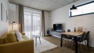 A modern living room with a sofa, a dining table with chairs, a TV, and a large window with curtains.