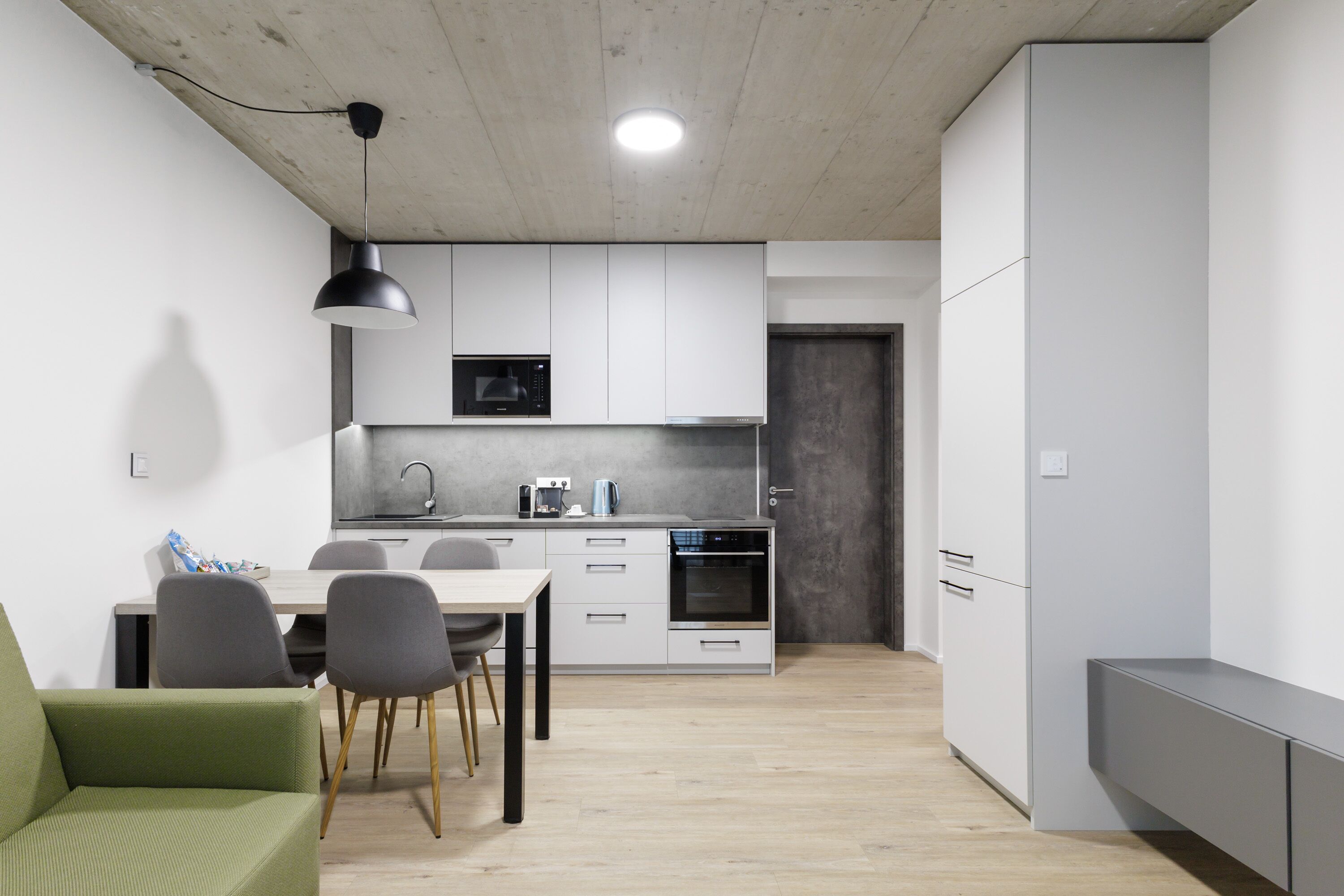 A modern kitchen with white cabinets, a microwave, and an oven. There is a dining table with chairs and a green sofa in the foreground.