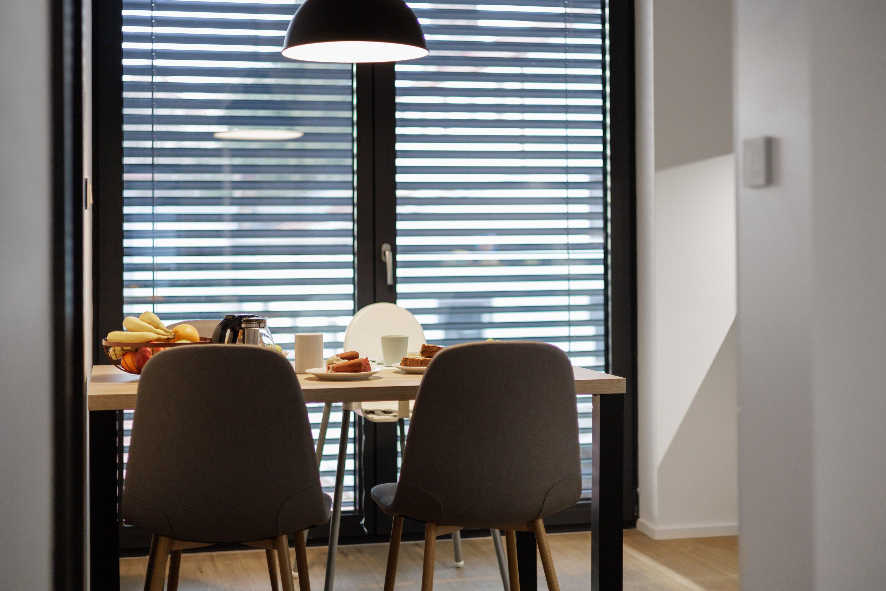 Un coin repas comprenant une table, deux chaises, une lampe et une fenêtre avec des stores.