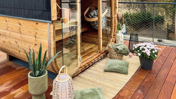 Een knus houten vakantiehuisje met een glazen deur, een klein zithoekje binnen en een houten terras met planten en een lantaarn.
