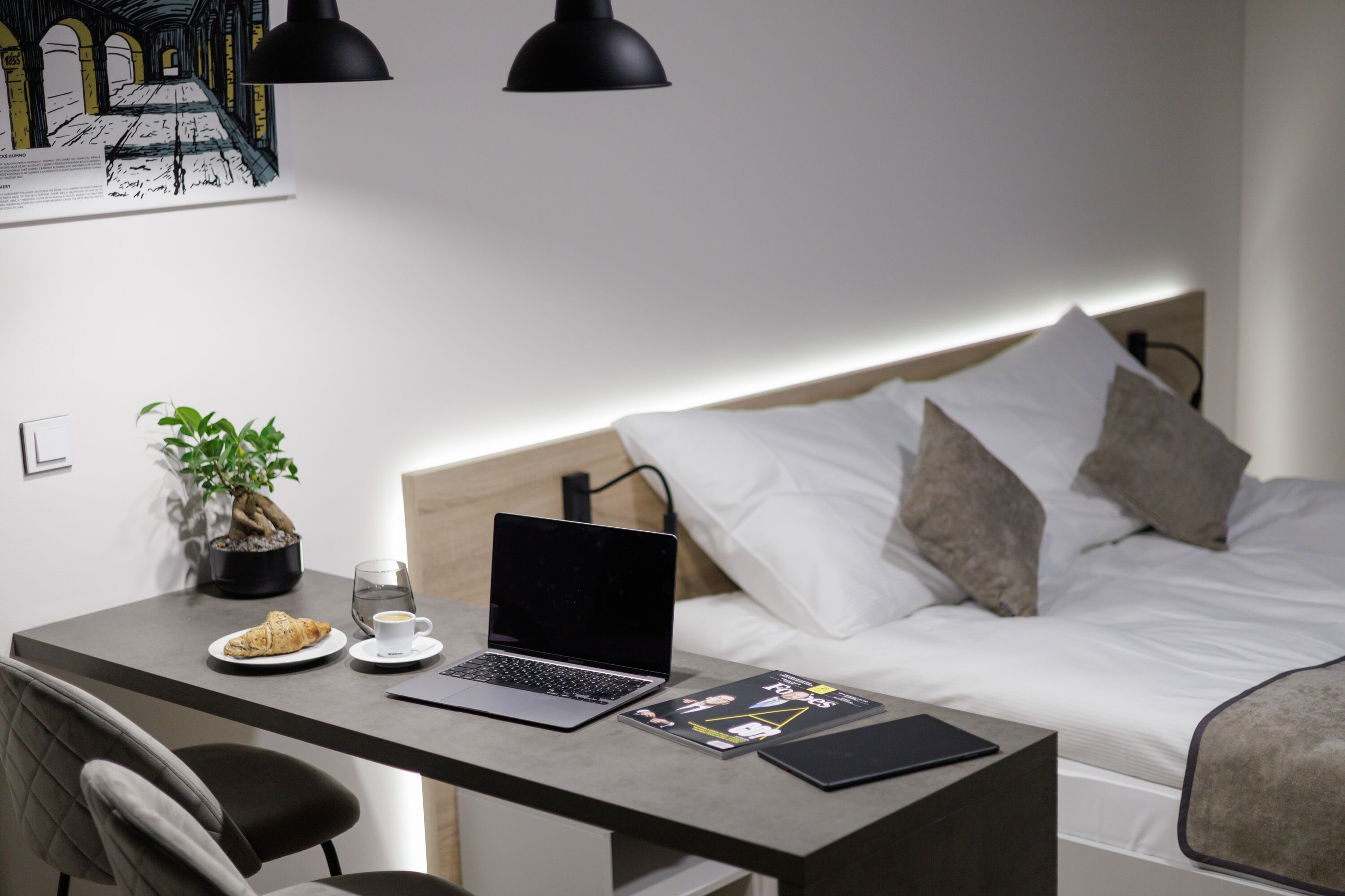 A modern bedroom with a bed, bedside table, laptop, and a small desk area with a chair and a potted plant.