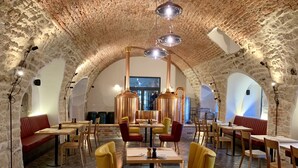 Een interieur met stenen muren, gewelfde plafonds en een koperen distillatie-apparaat in het midden. Er staan tafels en stoelen rondom de ruimte.