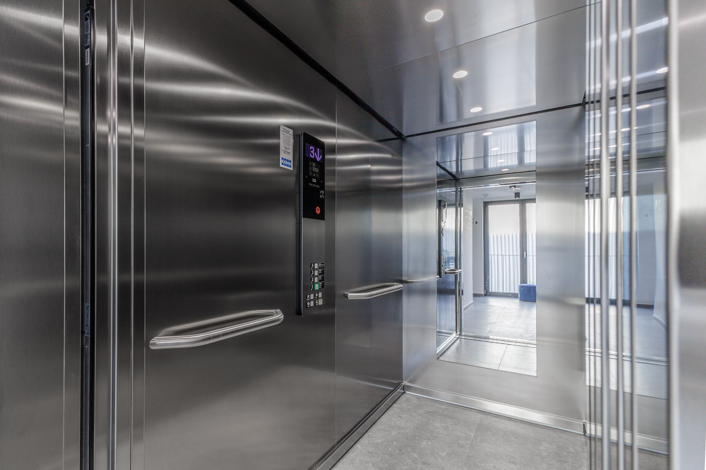 Un ascensor modern amb pantalla digital, parets metàl·liques i una porta de vidre transparent.