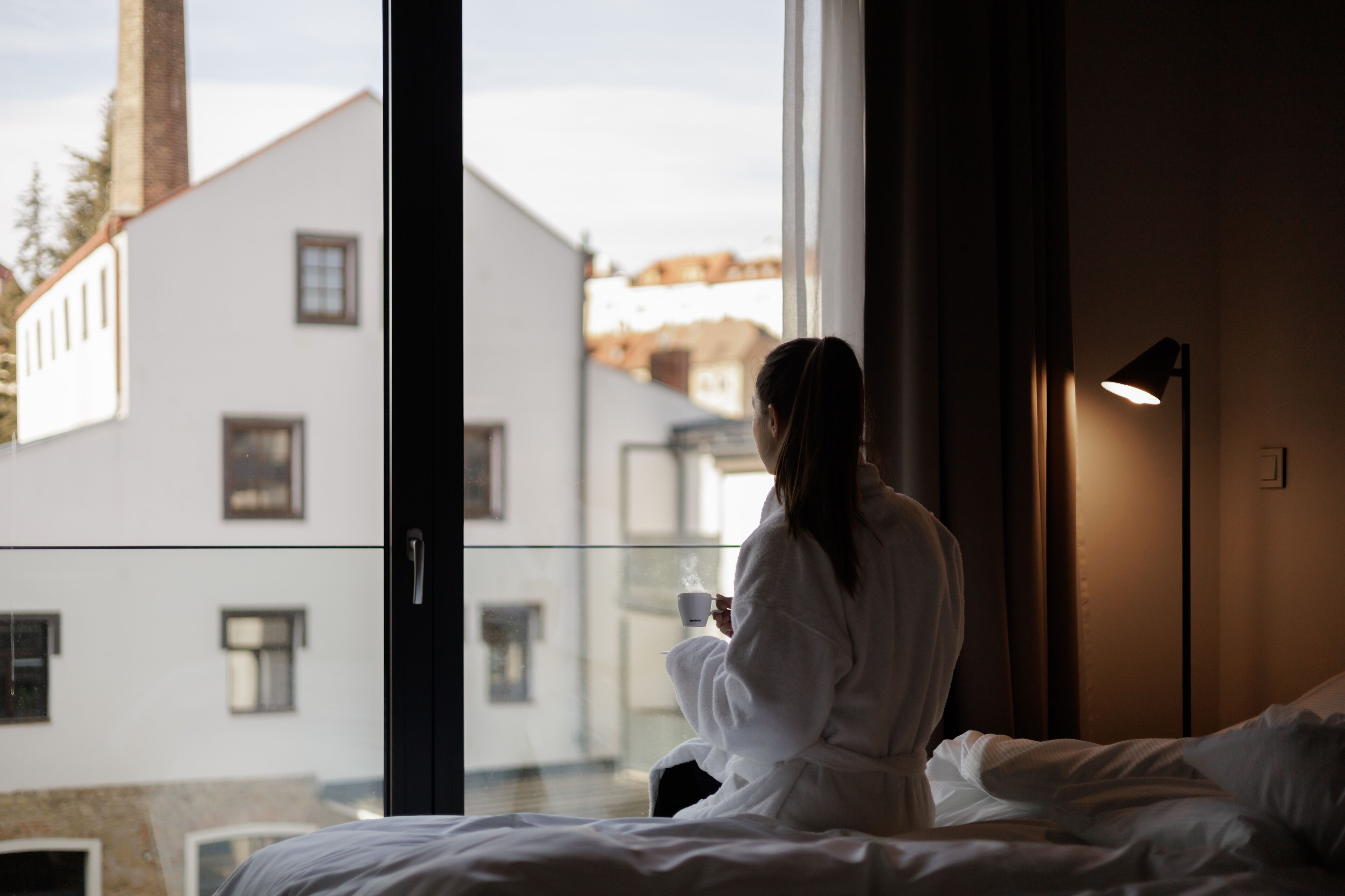 Une personne vêtue d’une longue robe blanche est assise sur un lit près d’une fenêtre, d’où l’on aperçoit des bâtiments à l’extérieur.