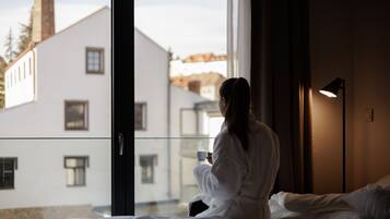 Une personne vêtue d’une longue robe blanche est assise sur un lit près d’une fenêtre, d’où l’on aperçoit des bâtiments à l’extérieur.