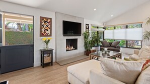 Ein modernes Wohnzimmer mit Kamin, Flachbildfernseher und großen Fenstern mit Blick auf Grünflächen.