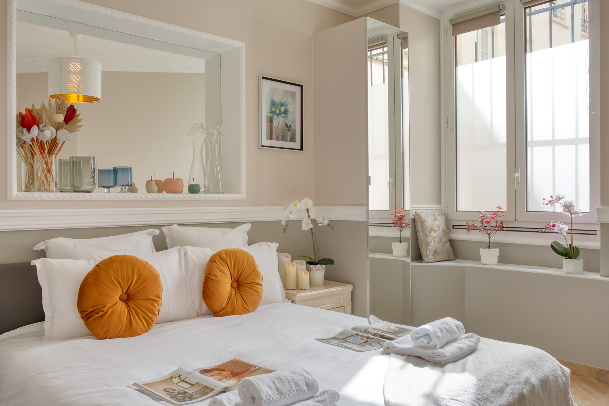 A neatly arranged hotel room with a large bed, two pillows, a bedside table with candles, a mirror, and a window with a view of the outside.