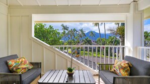 A balcony with wicker furniture, a small table with a plant, and a view of palm trees and mountains. - Liho Kai 4 Bedroom Home by RedAwning (Princeville)