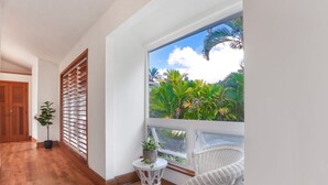 A room with a large window, a white wicker chair, a small round table with a plant, and a wooden door. - Liho Kai 4 Bedroom Home by RedAwning (Princeville)