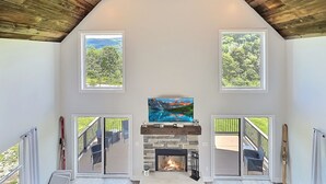 A spacious living room with a stone fireplace, two windows with a view of greenery, and a mounted painting above the fireplace. - New Build • Ellicottville Ski Chalet • Sleeps 8 (Ellicottville)
