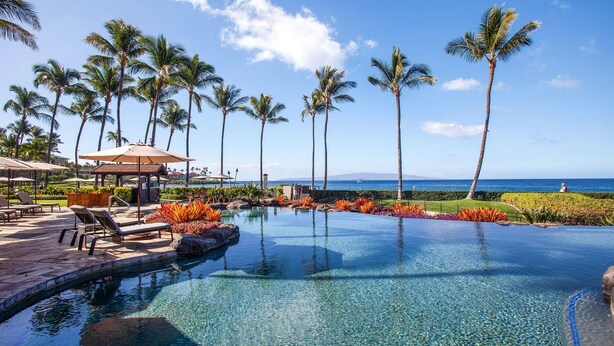 A clear blue pool surrounded by palm trees and lounge chairs, with a view of the ocean and a distant mountain. - Wailea Beach Villas M312 3 Bedroom Condo (Kihei)