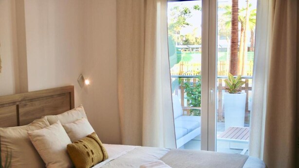 Quarto de hotel com cama, almofadas e vista para uma varanda com plantas e um deck de madeira.