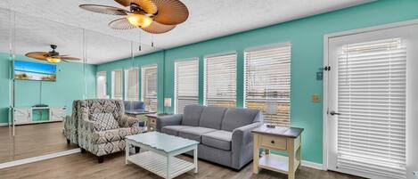 Una sala de estar con un sofá gris, un sillón estampado y una mesa de centro. Hay varias ventanas con persianas, un ventilador de techo y una puerta con ventanal.