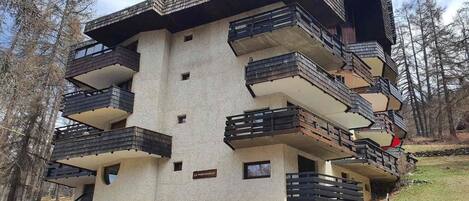 A multi-story building with balconies, a sign indicating 'Le Parcours,' and a natural surrounding.