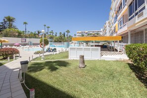 A pool area with lounge chairs and umbrellas, a small grassy area with a tree stump, and a building with balconies and windows. - Complejo Aloe - Pool - close to Maspalomas Beach (San Bartolomé de Tirajana)