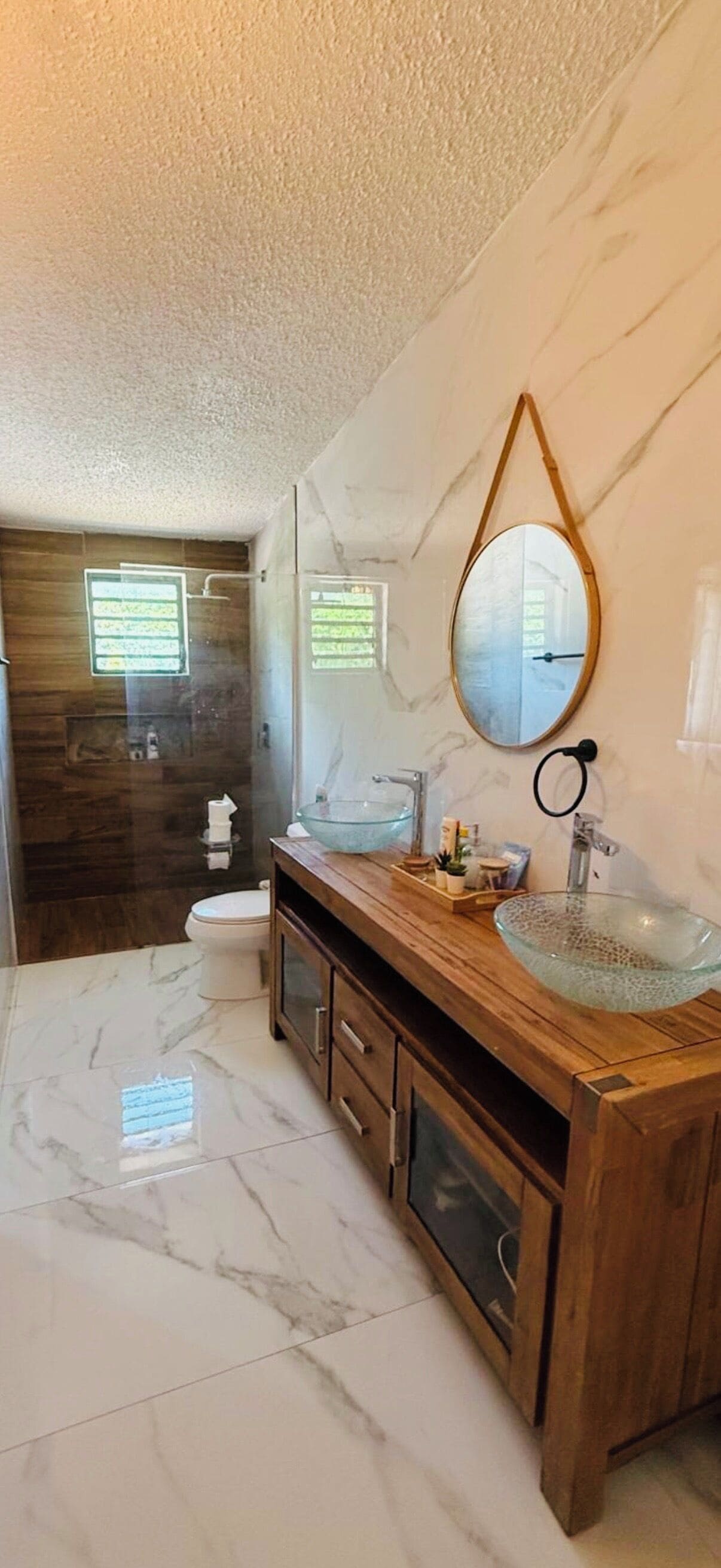 Un baño con tocador de madera, lavabo de vidrio, espejo grande y piso de mármol.