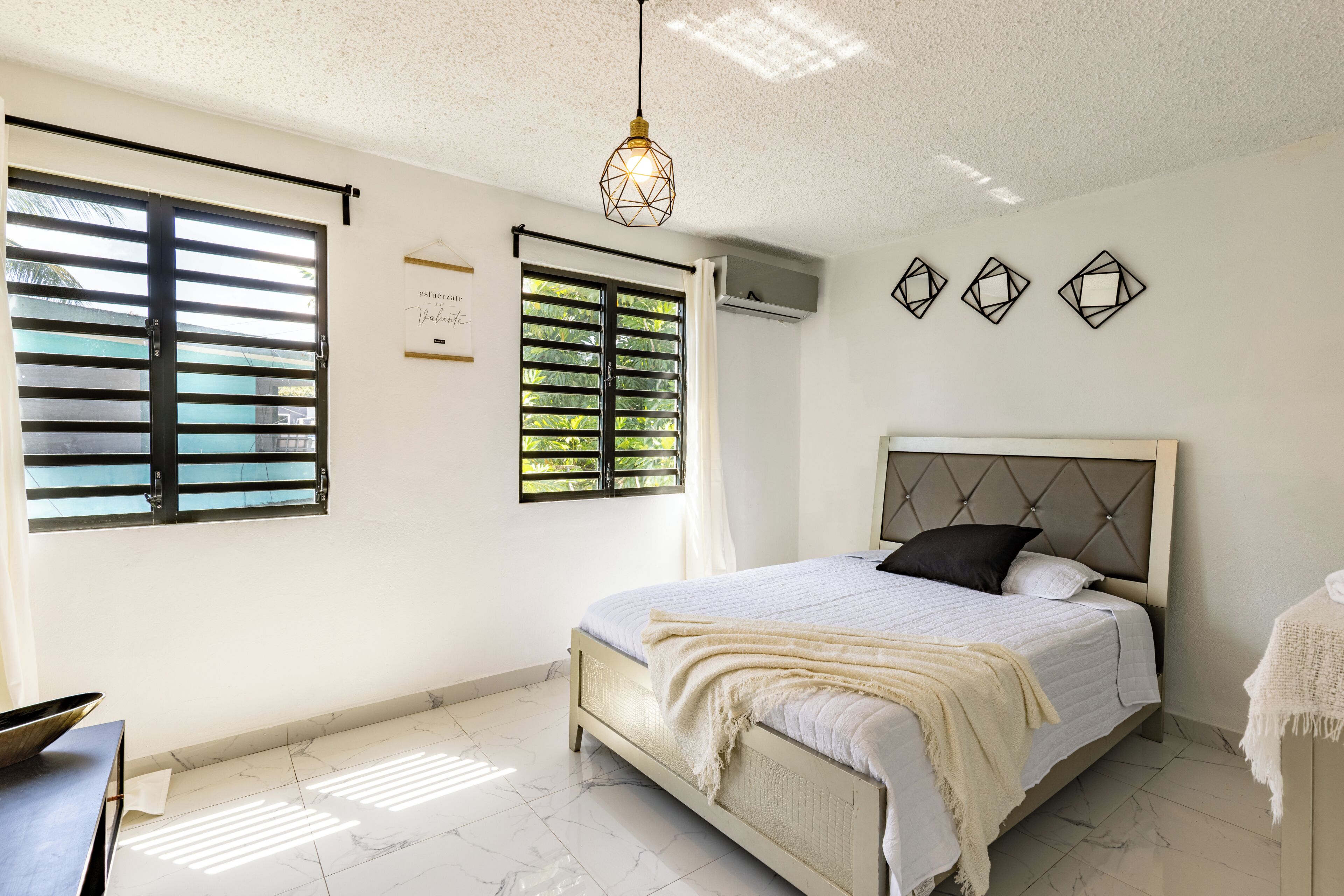 Une chambre à coucher avec un lit, une fenêtre avec des stores, un luminaire suspendu et un climatiseur fixé au mur.