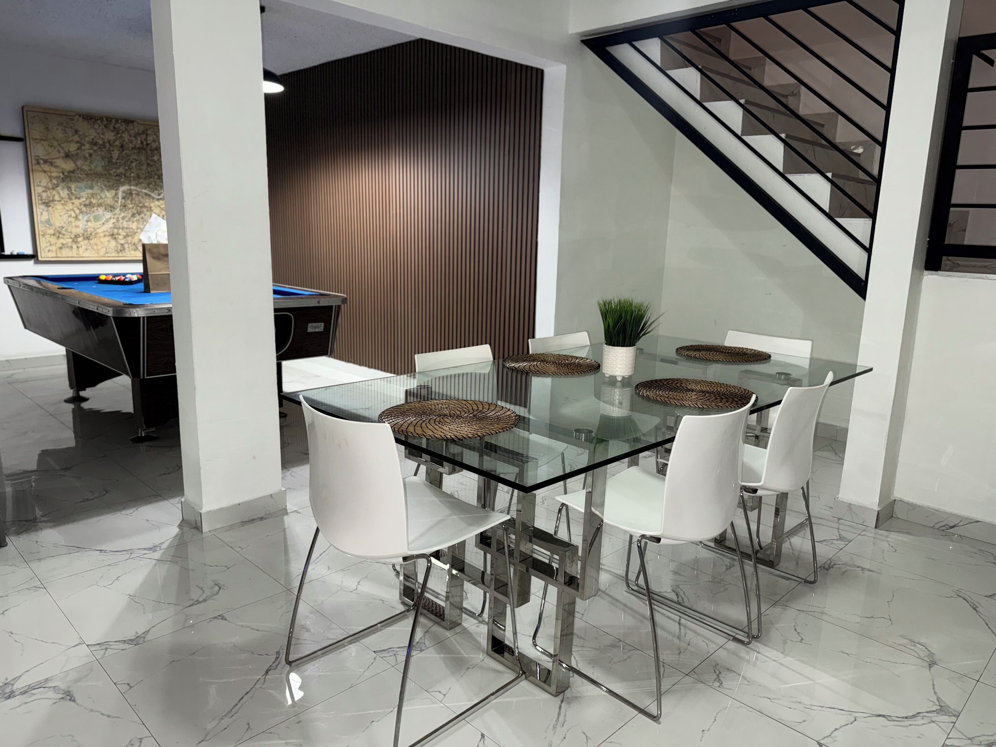Un espace repas moderne avec une table en verre entourée de chaises blanches, une table de billard à l’arrière-plan et un escalier sur la droite.