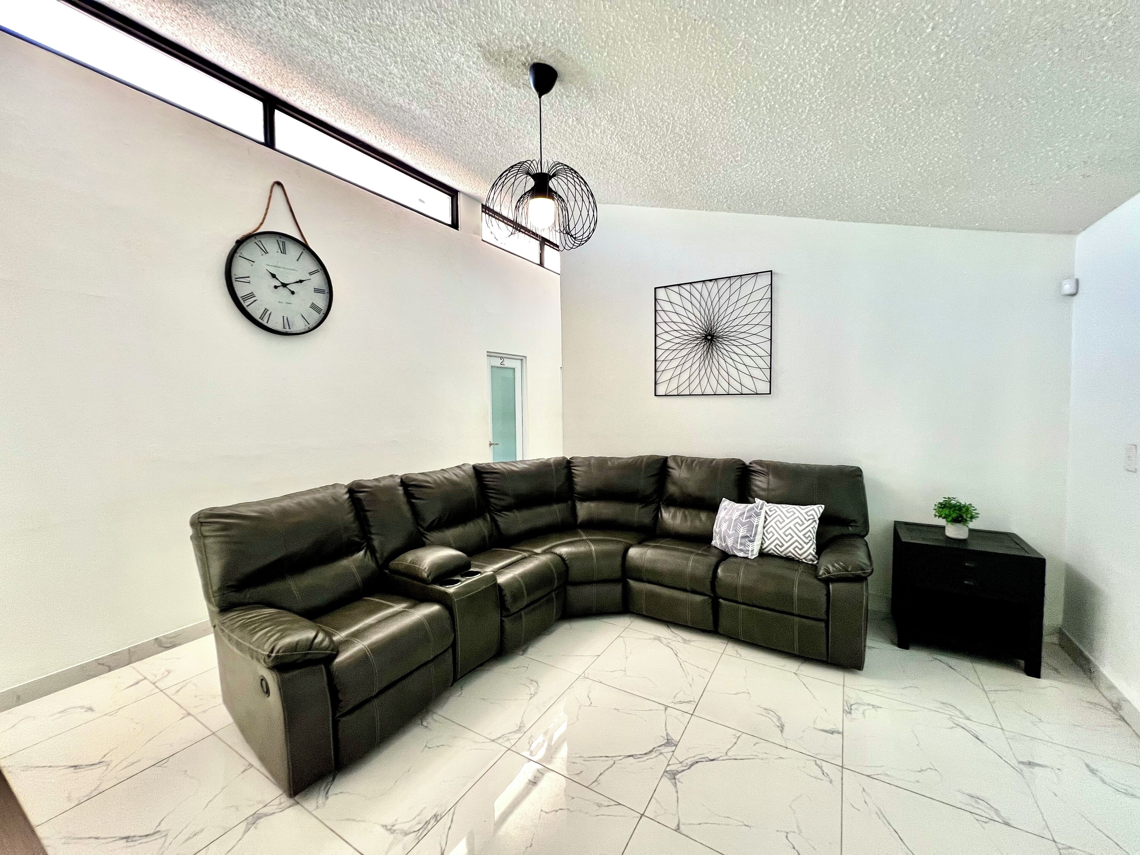 Ein modernes Wohnzimmer mit einem Ecksofa, einer Wanduhr, einem geometrischen Wandbild und einer Deckenleuchte.