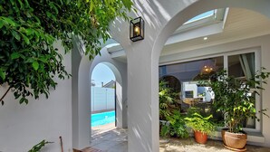 Uma área de varanda coberta com piscina, plantas em vasos e vista para o quintal.