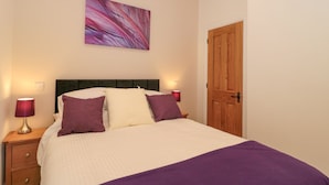 A neatly made bed with a white and purple bedspread, two purple pillows, and a headboard. There is a wooden nightstand with a lamp and a wall-mounted light. A wooden door is visible on the right. - The Nest (Bridgwater)