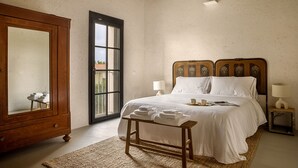 Een slaapkamer met een grote houten garderobe, een bed met witte lakens, een klein nachtkastje met een lamp en een groot raam met uitzicht op het omliggende landschap.