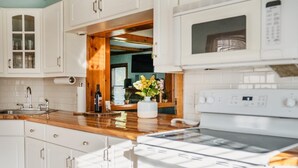 Une cuisine avec des meubles blancs, un plan de travail en bois, un four encastré et une fenêtre avec des stores.