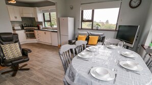 Un comedor con una mesa redonda preparada para seis personas, rodeada de sillas. Se aprecia una cocina al fondo con armarios blancos y un refrigerador.