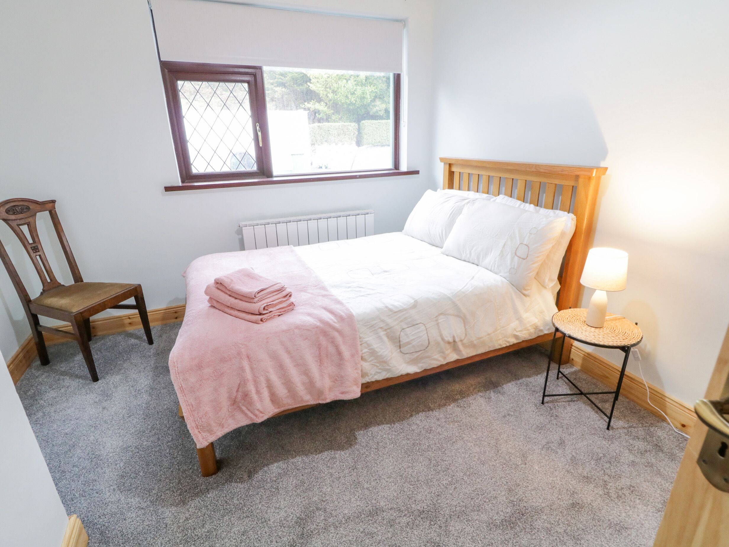 A bedroom with a bed, a chair, a bedside table with a lamp, a window, and a radiator.