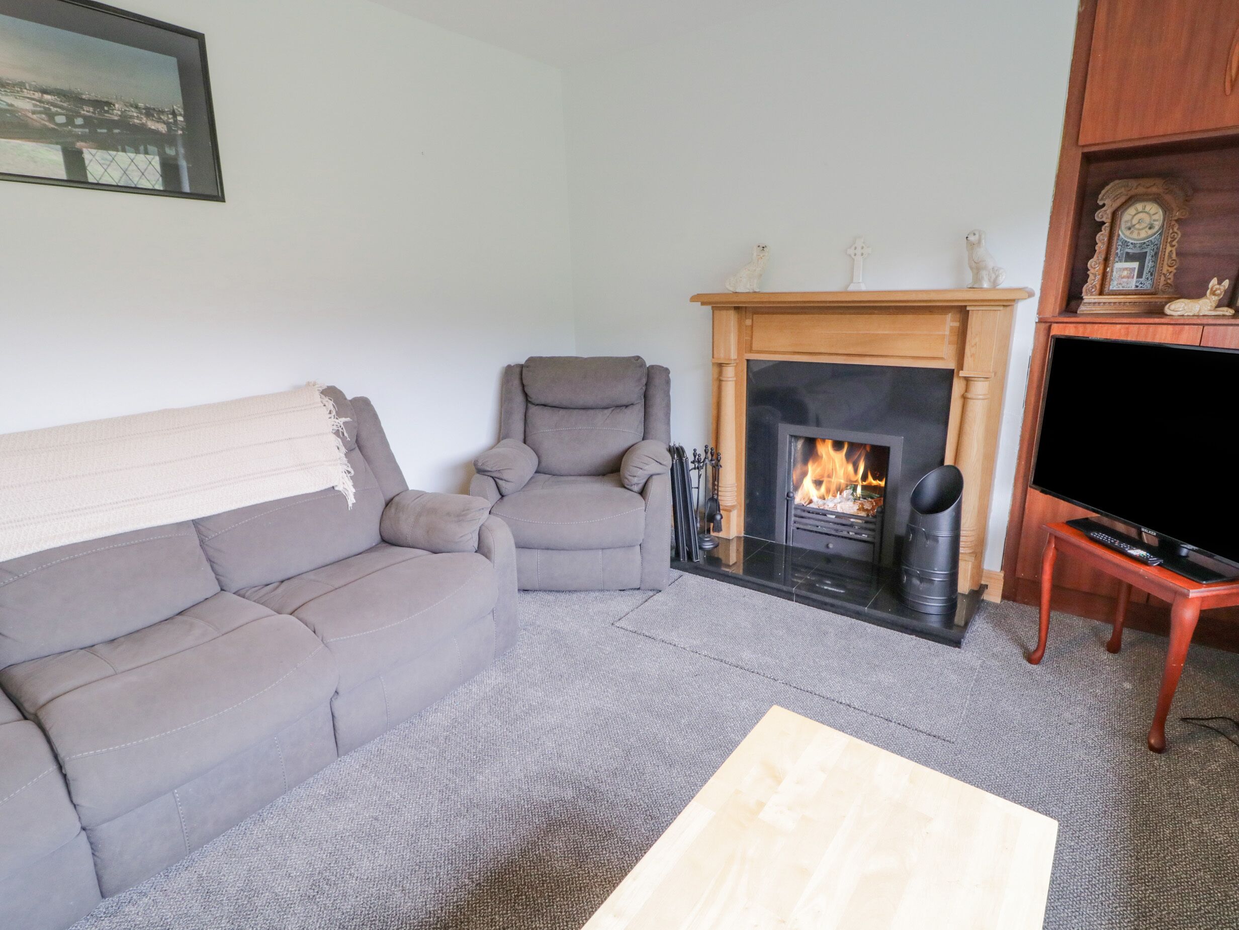 A living room with a grey sofa, a grey armchair, a fireplace with a fire, a television, and a wooden cabinet with a clock.