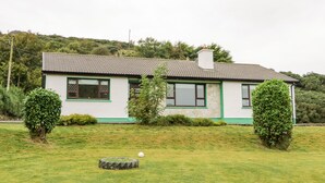 Una casa de dos pisos con techo de tejas, paredes blancas y detalles en verde. Tiene dos ventanas con contraventanas, una chimenea y un pequeño jardín con césped y dos arbustos.