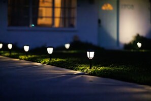 A pathway lined with evenly spaced outdoor lamps, leading to a building with a visible window and a door.