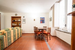 Una sala de estar con sofá, una mesa de comedor con sillas, una estantería y un ventanal con cortinas.
