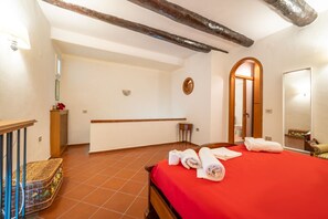 A hotel room with a red bedspread, wooden beams on the ceiling, a small table with towels, and a mirror on the wall. - Il Nido - One Bedroom Apartment, Sleeps 3 (Naples)