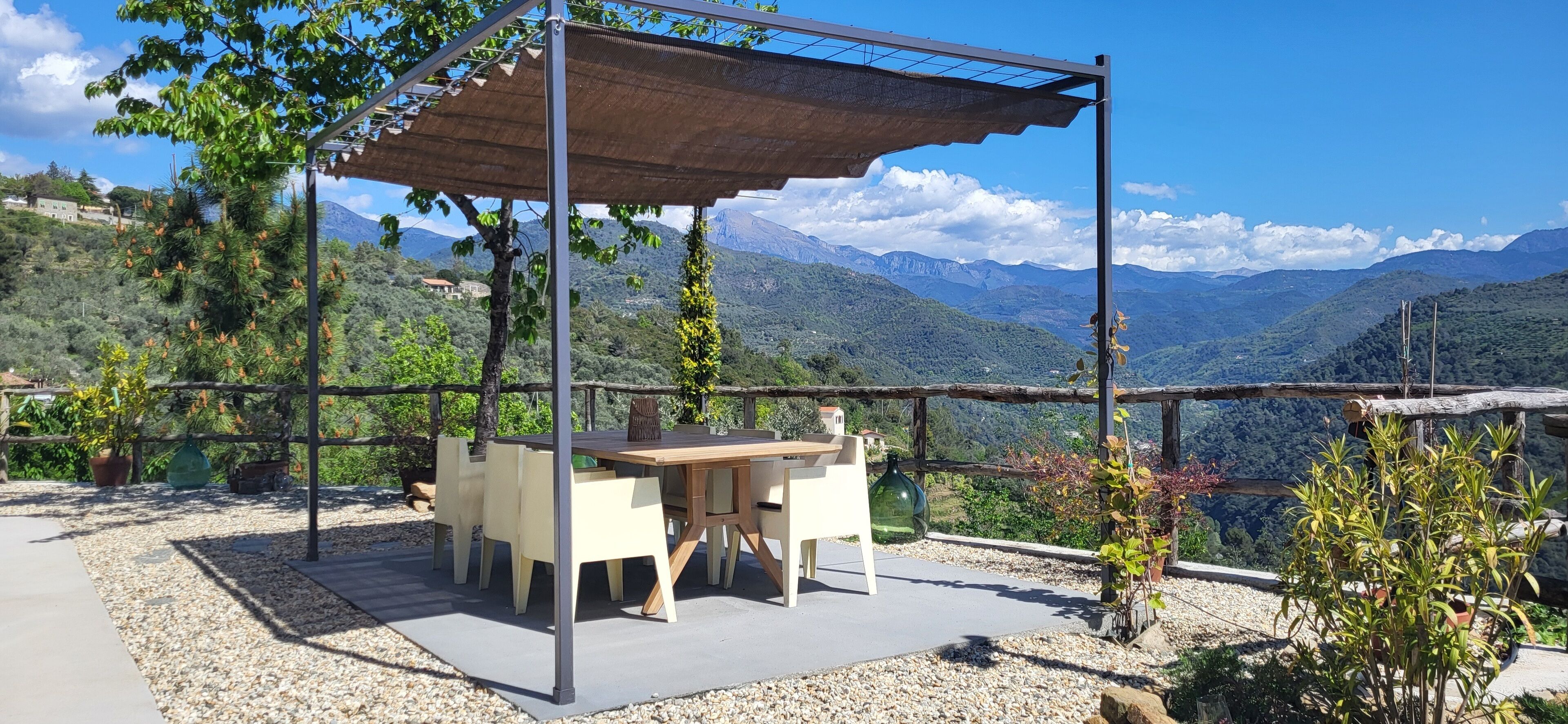 Un espacio de estar al aire libre con una mesa y sillas bajo una pérgola, rodeado de grava y plantas en macetas, con vistas a un paisaje montañoso.