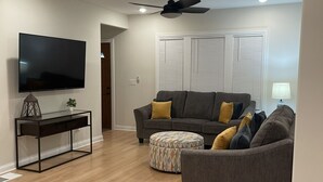 Ein Wohnzimmer mit Flachbildfernseher, einem schwarzen Konsolentisch, einem grauen Sofa und einem gemusterten Ottomanen.