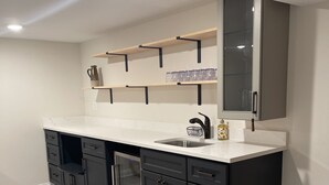 Eine moderne Küche mit dunklen Schränken, einer weißen Arbeitsplatte und einer Spritzschutzwand. Darüber befinden sich Holzregale und zur Rechten ein Glastisch.
