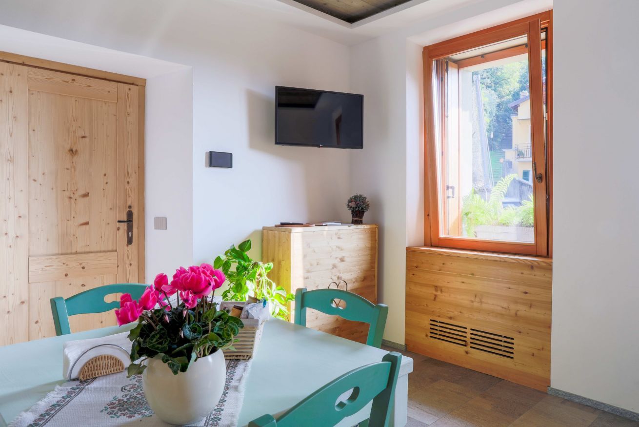 Una habitación con puerta de madera, televisor montado en la pared, ventana con vistas a follaje, mesa con sillas y planta en maceta con flores rosadas.